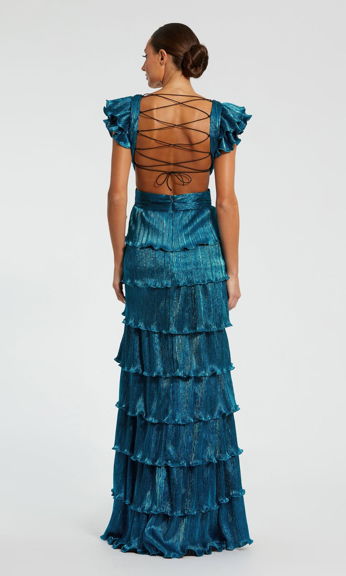 A woman faces away from the camera in a teal Mac Duggal Open-Back Tiered Formal Dress 26740, featuring ruffles, a floor-length v-neck design, and an elegant lace-up open back, set against a plain background.